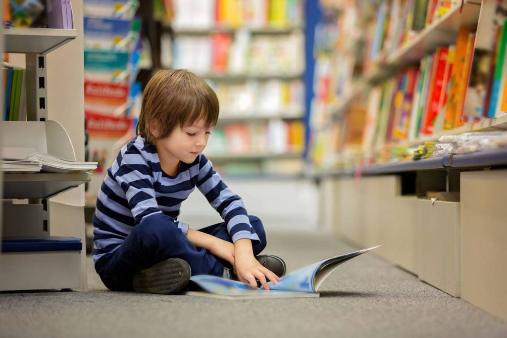 A young child sitting on a library floor, absorbed in reading a picture book among shelves of books