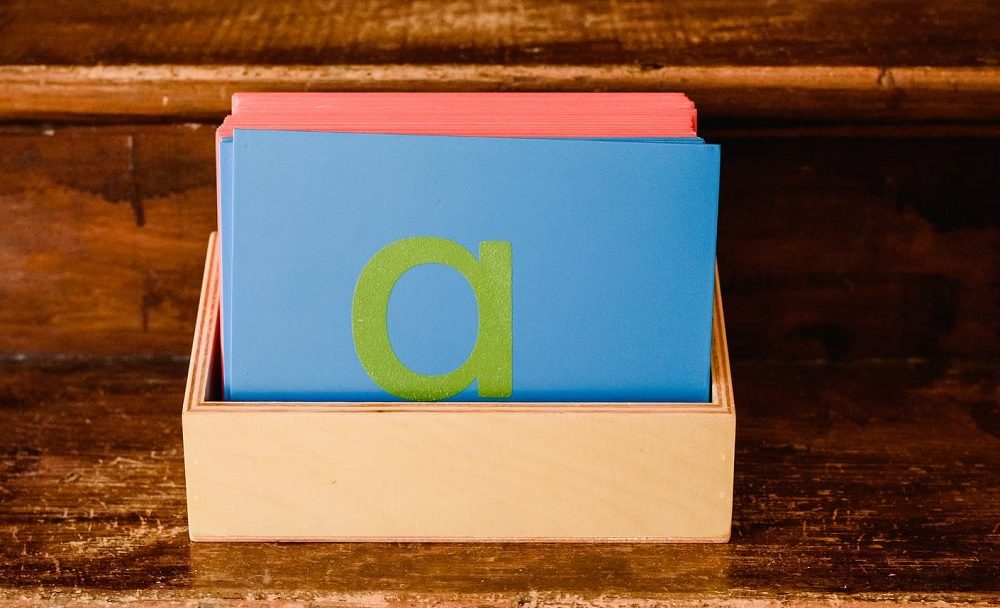 A Montessori Sandpaper Letter, a green lowercase 'a' on a blue board, standing in its wooden storage box