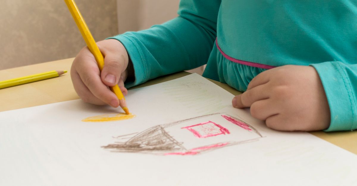 A young child holding a colored pencil, tracing and filling in a drawn shape on paper as handwriting preparation