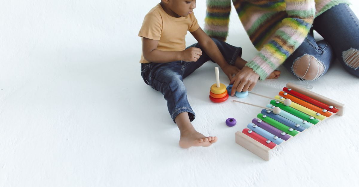 A young child and an adult sitting on the floor with a colorful wooden xylophone and a stacking-ring toy, exploring musical sounds