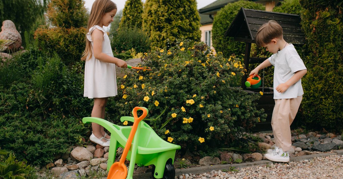 Two children playing outdoors in a garden, one watering flowering shrubs while the other pushes a small wheelbarrow
