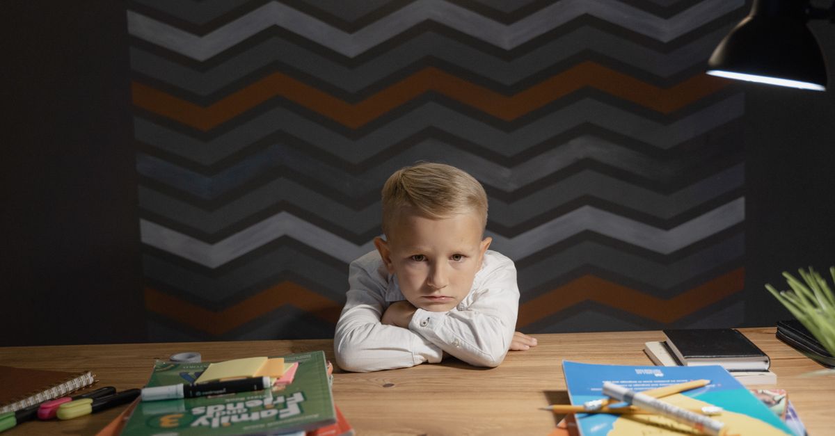 A school-age boy resting at a desk covered with books and school supplies, looking thoughtful