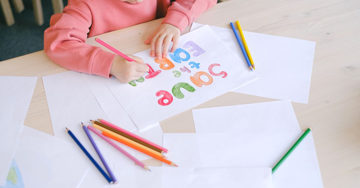 A child forming colorful letters on paper with a pencil, surrounded by colored pencils