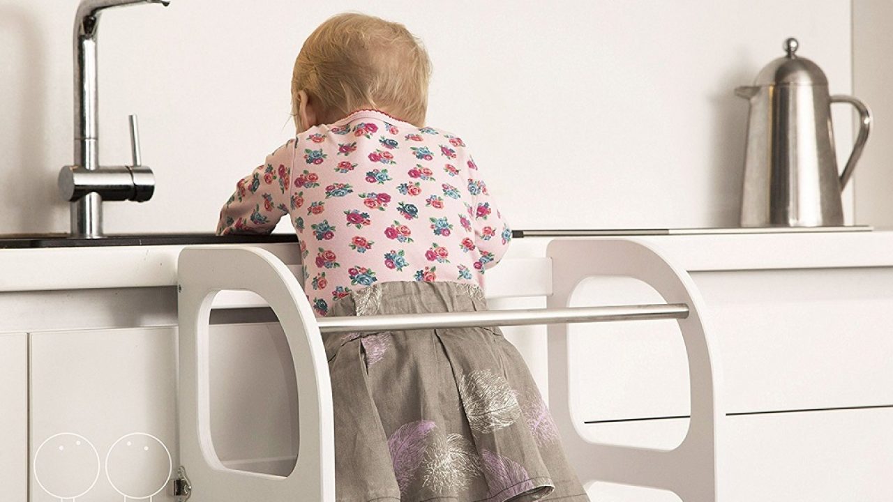 A young child standing safely in a wooden learning tower at a kitchen counter, participating in food preparation